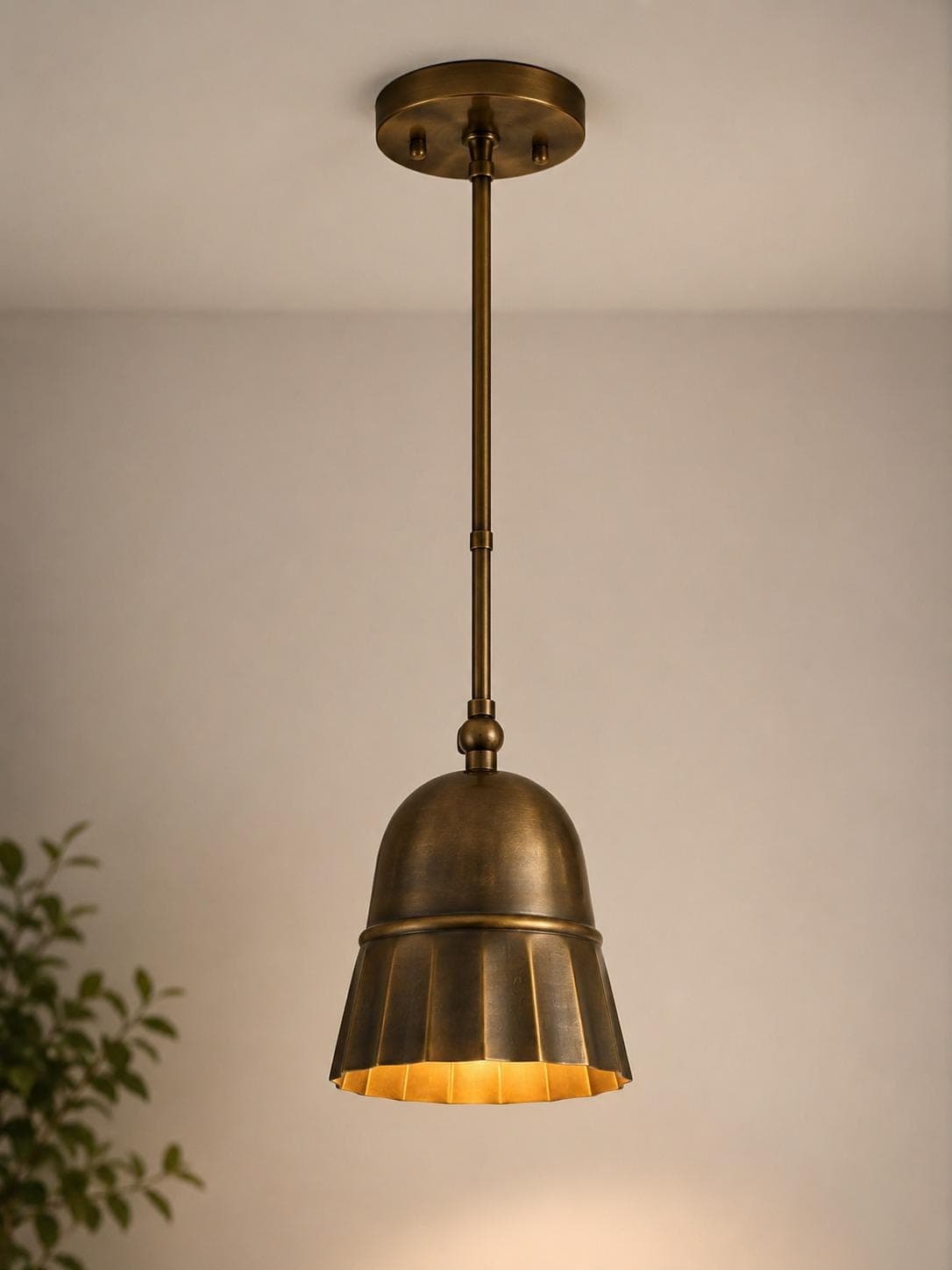 Antique brass pendant light with a domed top and fluted flared skirt hanging from a slim rod, lit warm against a pale wall