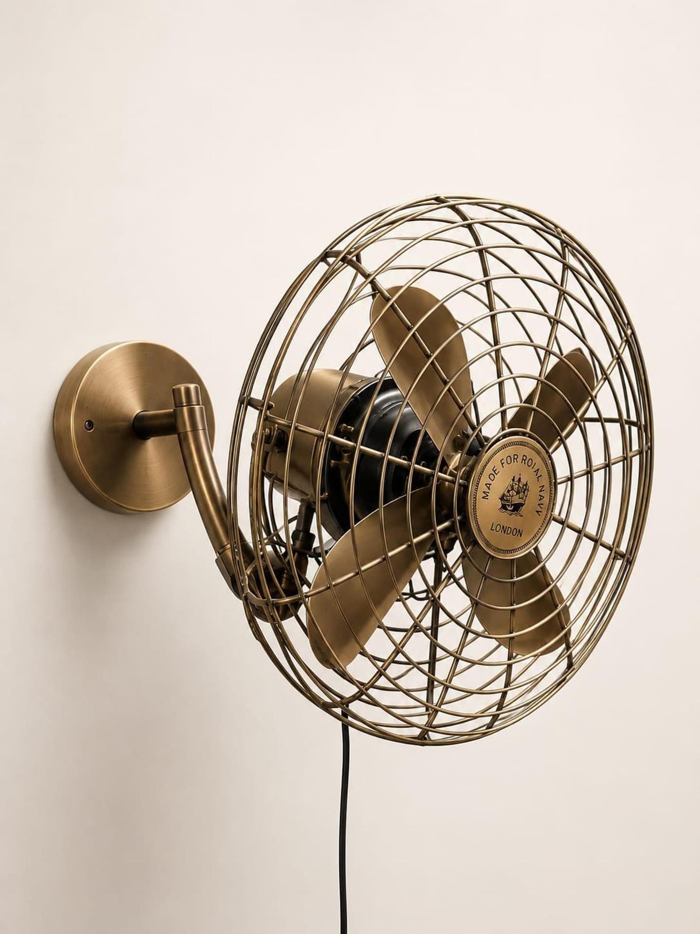 Brass wall-mounted oscillating fan with a fine brass cage and nautical hub badge on a gooseneck arm and round backplate