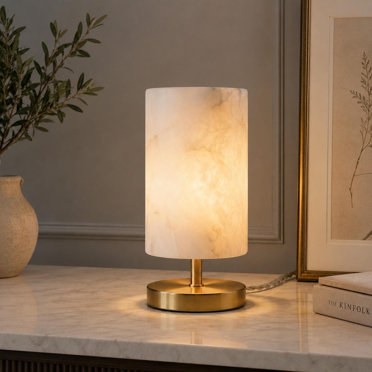 Alabaster cylinder table lamp lit on a marble console beside a framed print and a ceramic vase