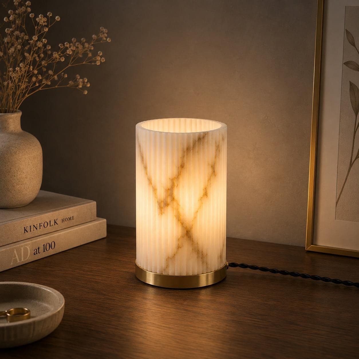 Fluted alabaster table lamp lit on a wooden nightstand beside stacked books and a framed print