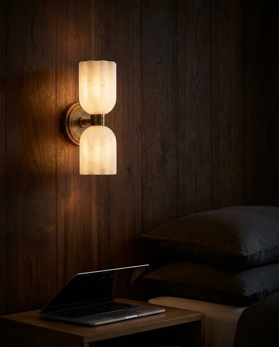 Fluted alabaster double-globe wall sconce glowing on a dark wood-paneled wall beside a nightstand and laptop
