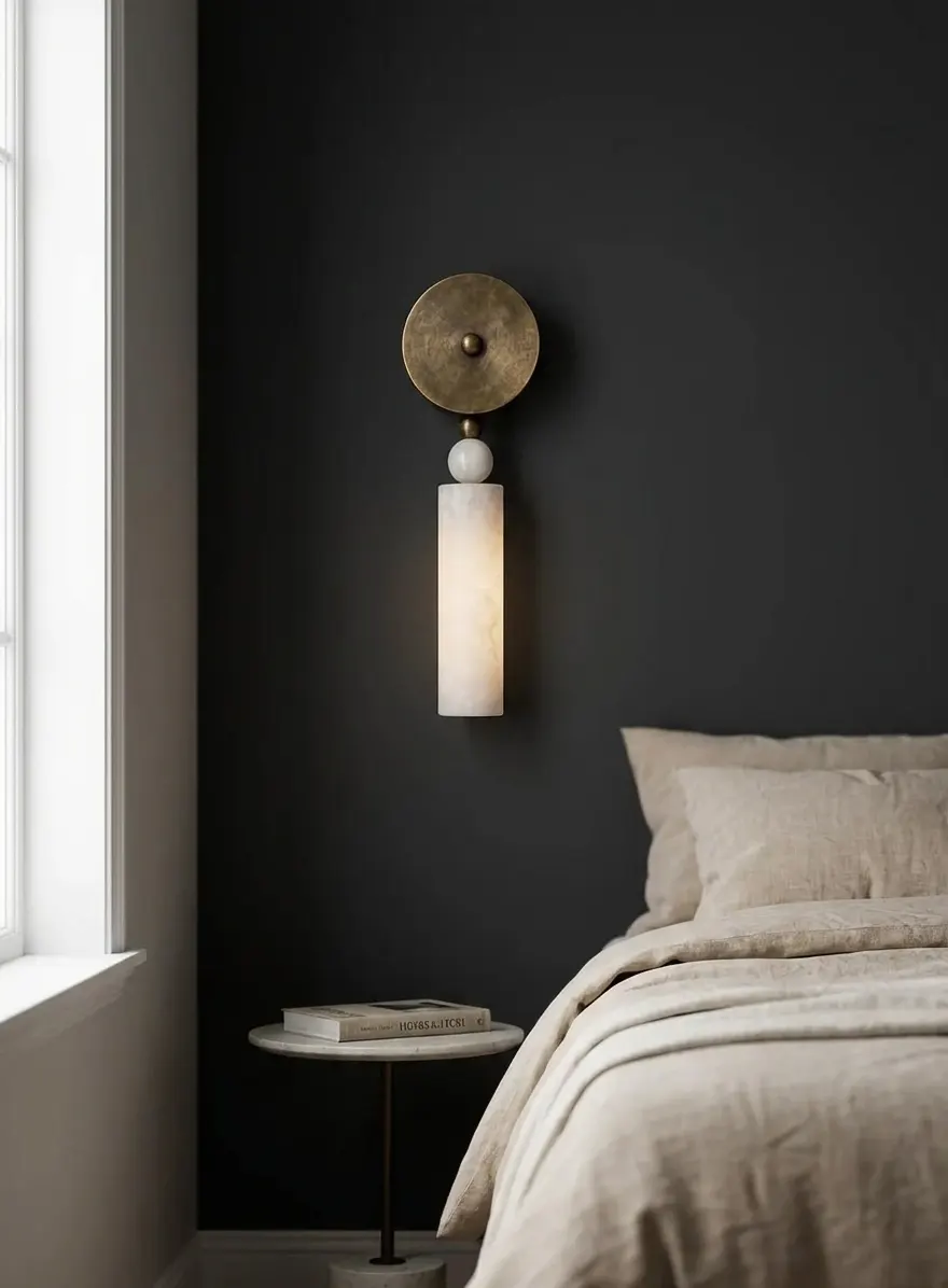Alabaster ball-and-cylinder pillar wall sconce glowing on a dark bedroom wall beside a nightstand