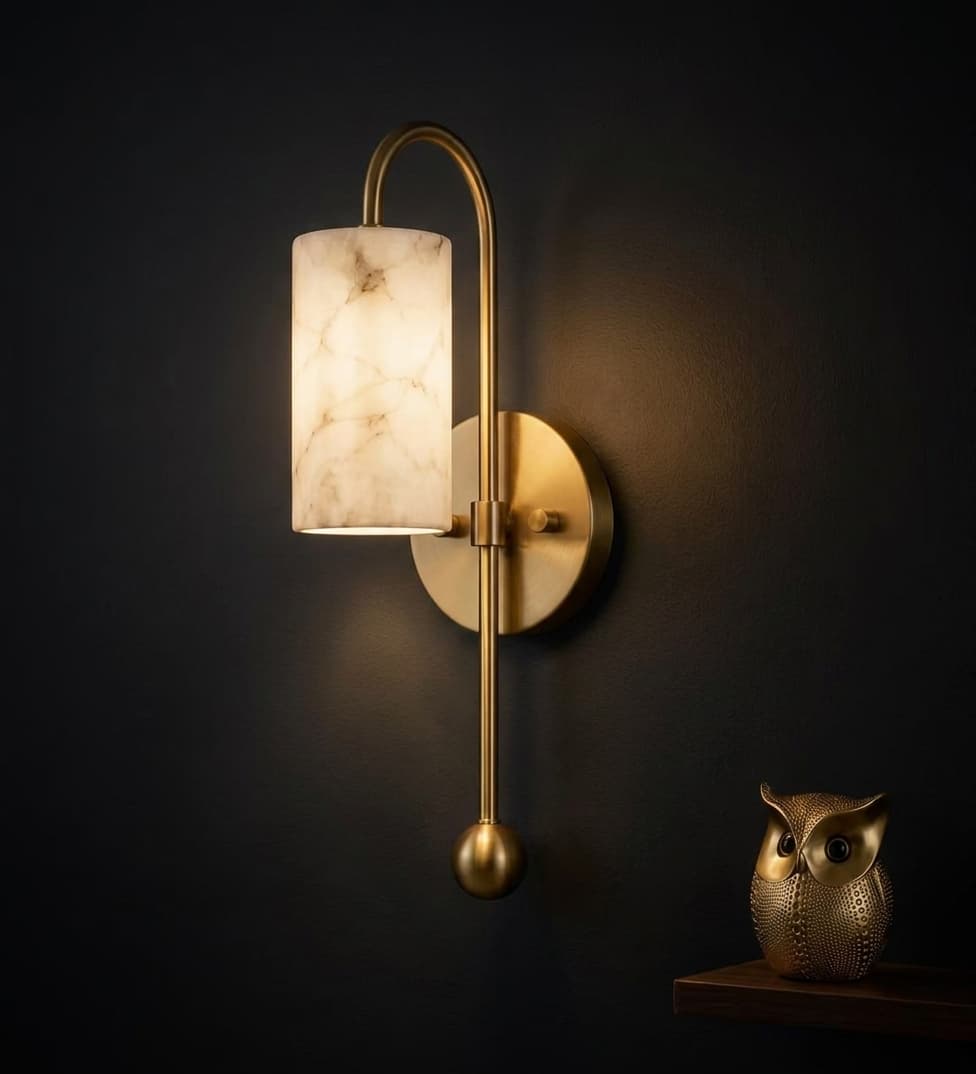 ArtLoka solid brass alabaster cylinder sconce, lit, on a dark navy wall beside a brass owl figurine
